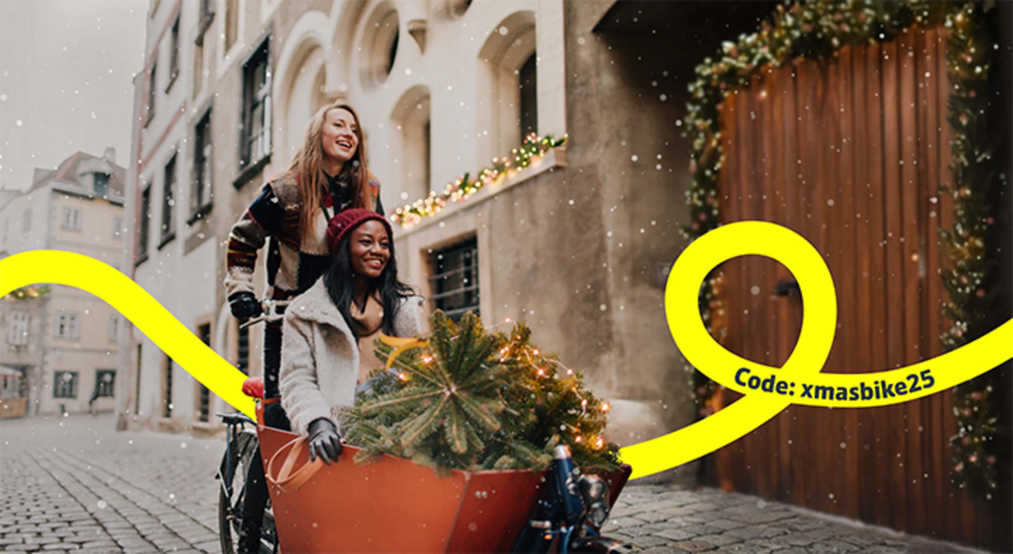 Zwei Frauen fahren mit einem Lastenrad und einem kleinen Weihnachtsbaum durch eine Stadt, in der Schnee fällt.