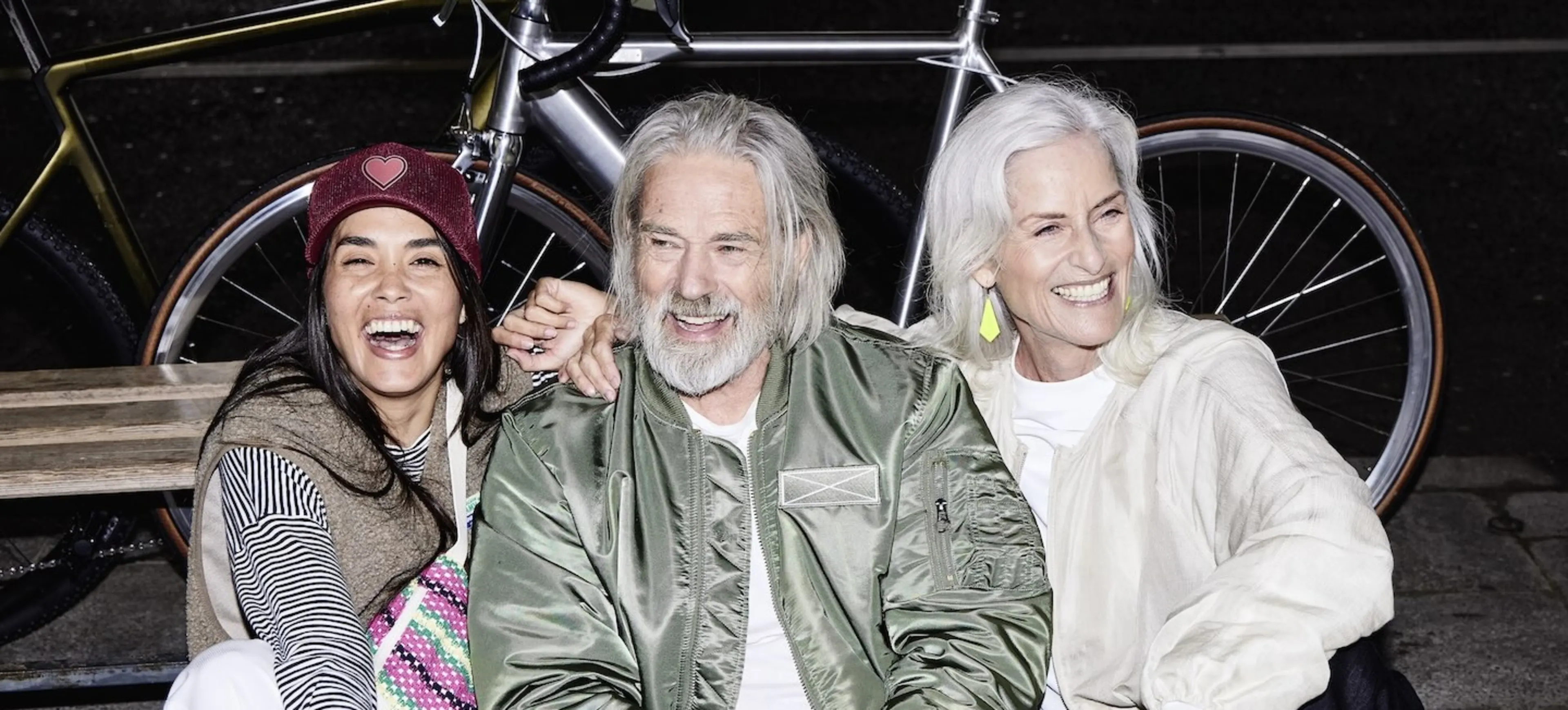 3 Personen sitzen lachend auf dem Gehsteig und hinter ihnen steht ein Fahrrad.