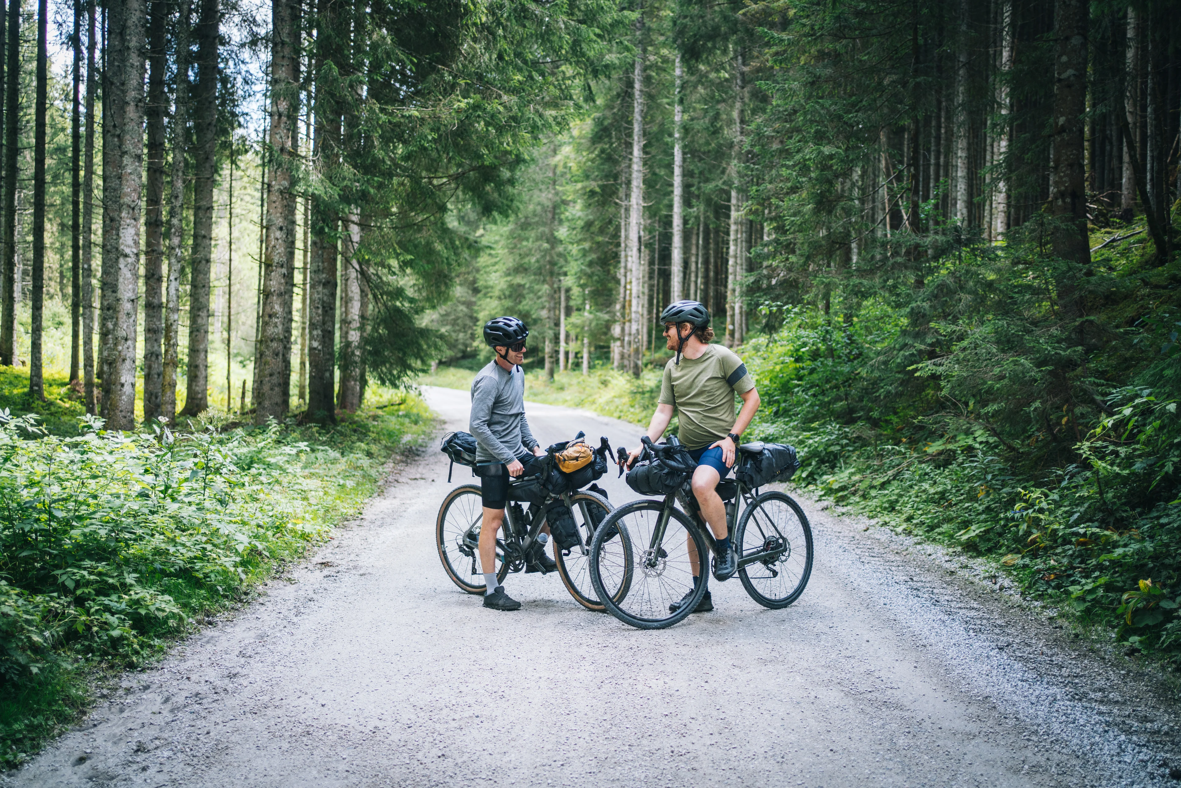 Zwei Bikepacker mit vollbepackten Gravelbikes machen eine Pause auf einem Waldweg und unterhalten sich.