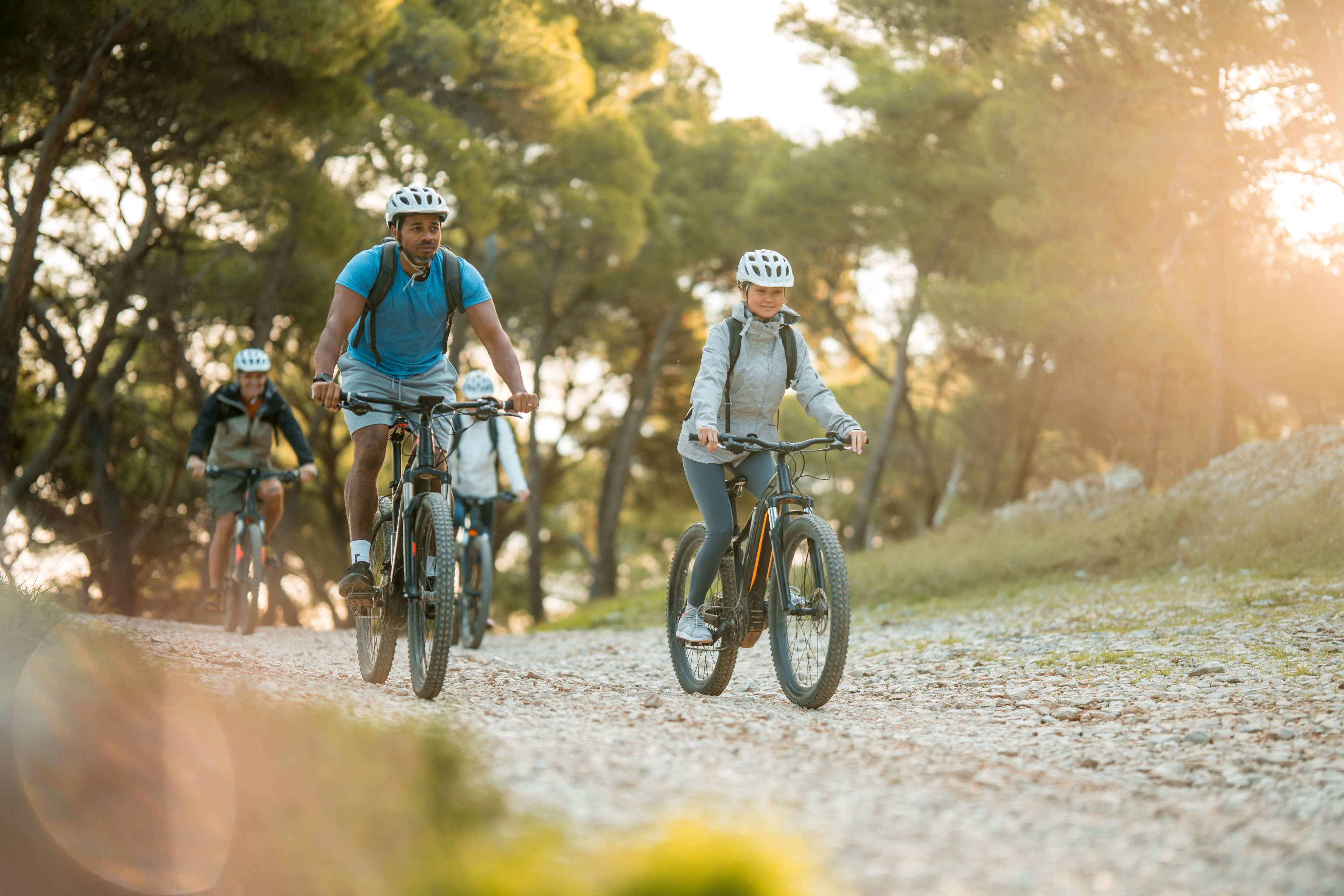 Vier Personen fahren mit Mountainbikes auf einem Schotterweg durch einen lichten Wald. Alle tragen weiße Fahrradhelme und Rucksäcke. Im Vordergrund sind zwei Radfahrer zu sehen – ein Mann im blauen T-Shirt und eine Frau in einer grauen Jacke. Im Hintergrund folgen zwei weitere Radfahrer. Die Szene ist in warmem Gegenlicht getaucht.