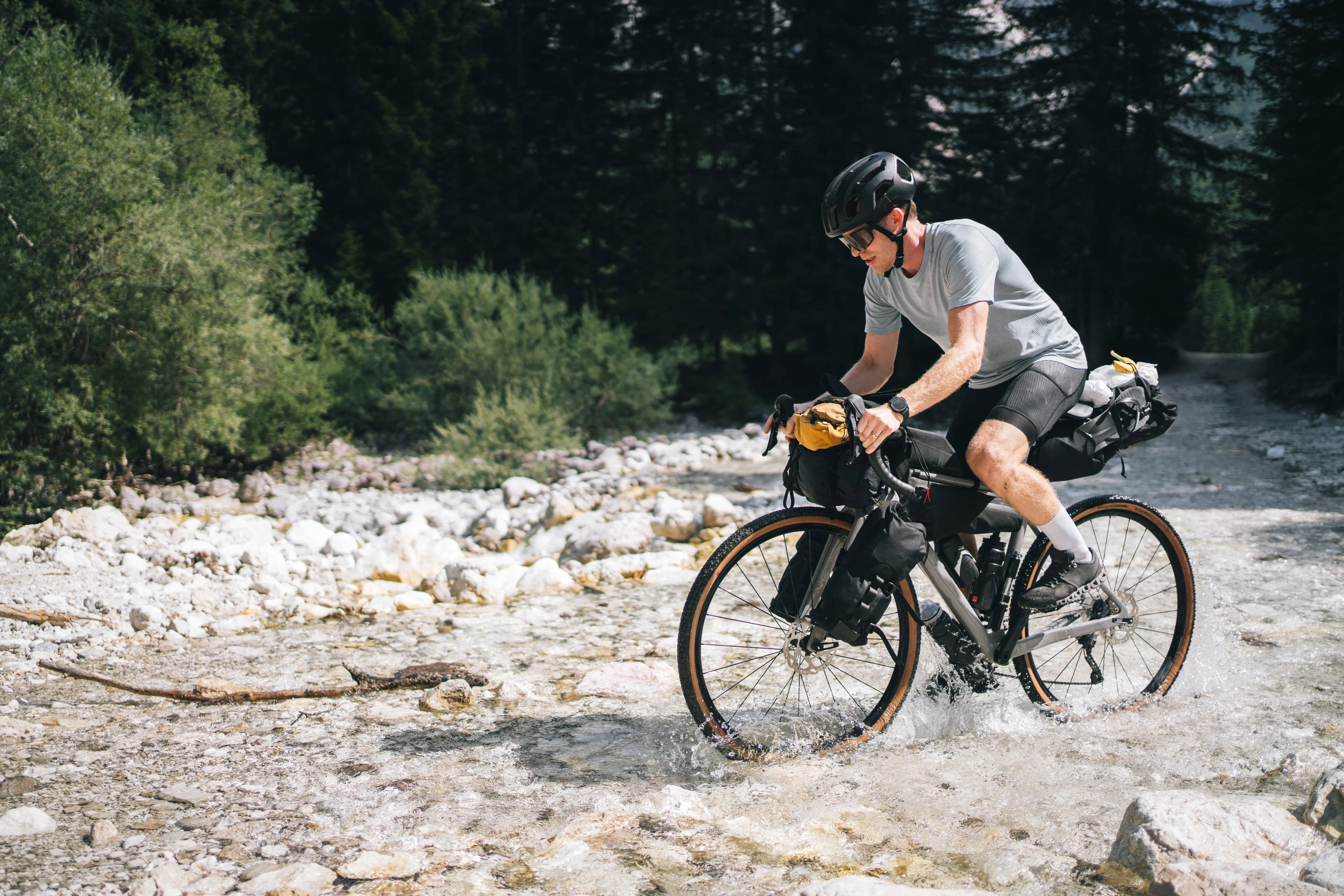 Ein Bikepacker mit schwarzem Helm und vollbepacktem Gravelbike durchquert einen flachen Gebirgsbach in felsiger Landschaft.