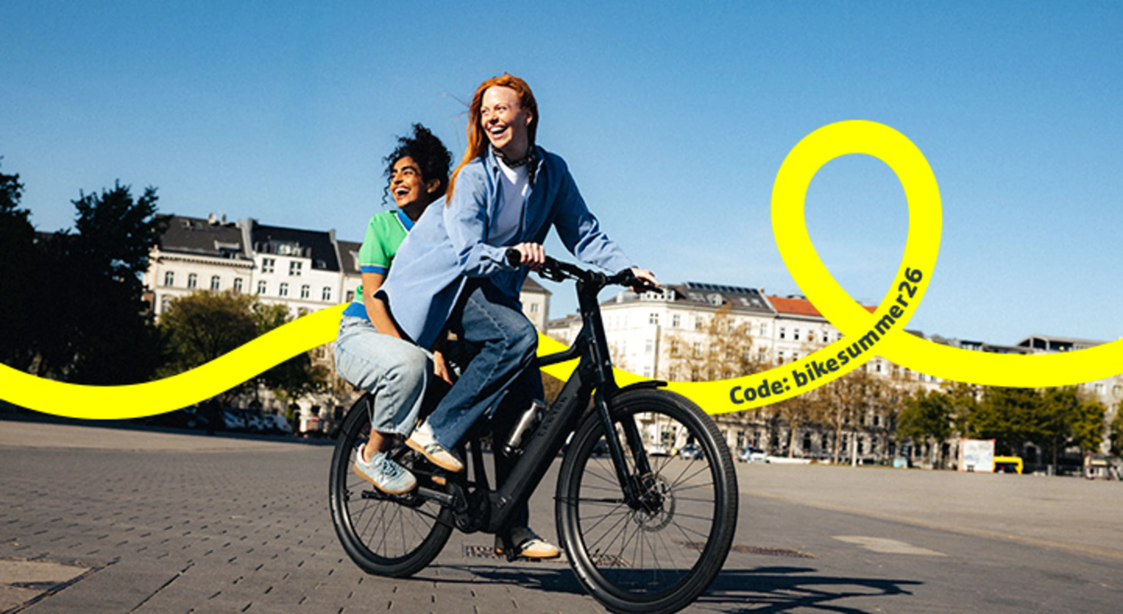 Zwei Frauen fahren an einem sommerlichen Tag gemeinsam auf einem E-Bike durch eine Stadt.