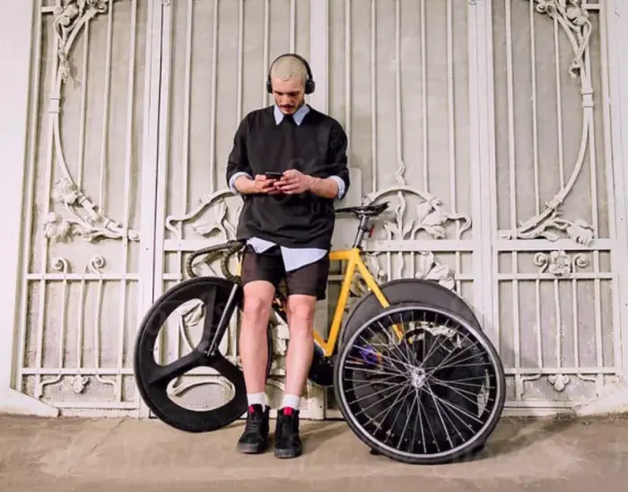 Ein Mann steht vor einem Tor. Er hat ein Handy in der Hand und Kopfhörer auf. Hinter ihm steht ein Rennrad. Er hat zudem einen Rucksack und ein zweites Laufrad dabei.
