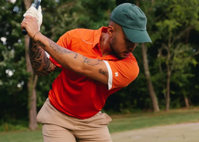 Male model wearing orange polo and khaki shorts, wearing green cap, swinging golf club