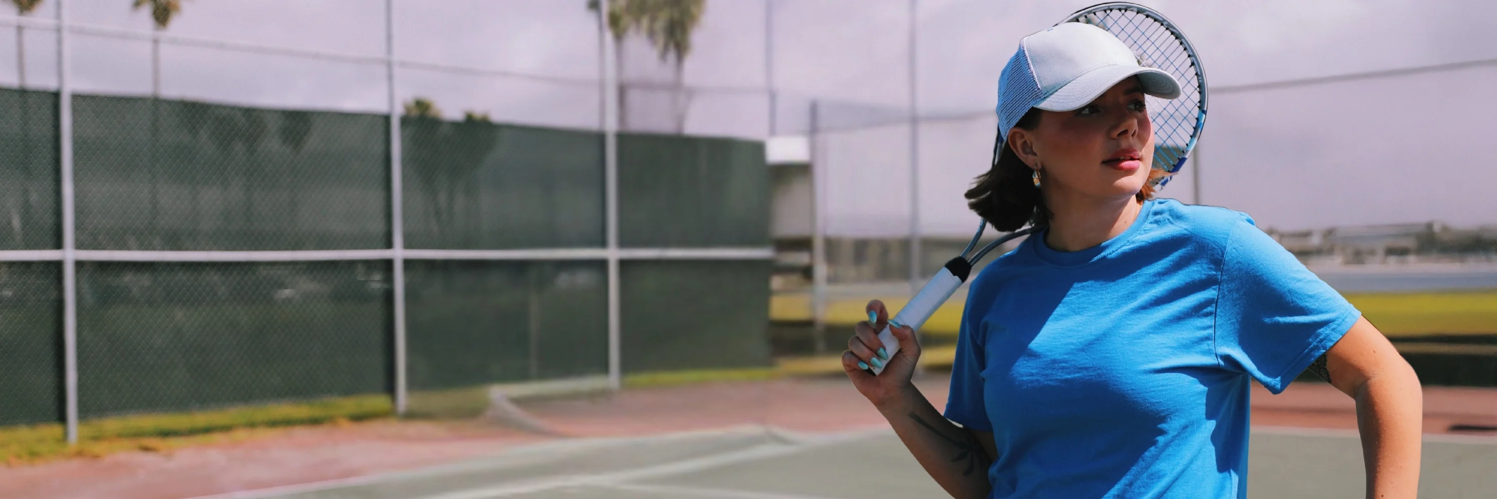 Female model wearing blue russell athletic t-shirt, pink skirt and cap holding a tennis racket at a tennis court