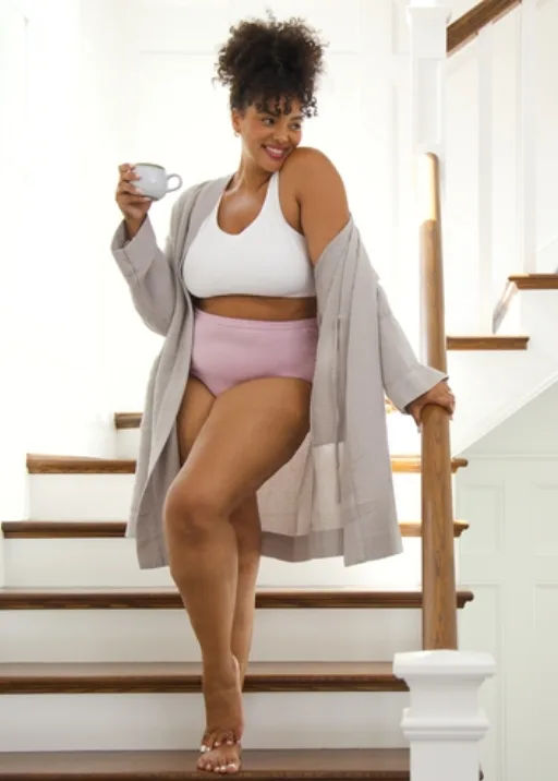 women wearing fruit of the loom pink underwear and white bra standing on a staircase with a cup of coffee. She also has khaki colored cardigan draped around her arms and shoulders.