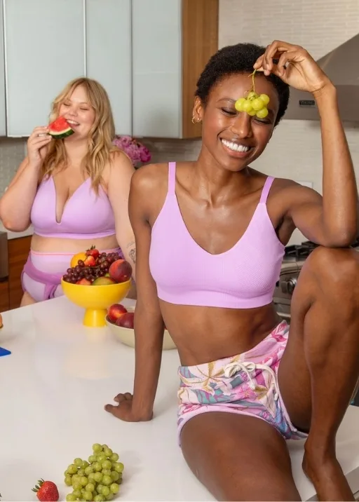 Two young women are in a kitchen with fruit all around them. The girl in the back is wearing a matching light purple bra and underwear while eating a slice of watermelon, the other is sitting on top of the island wearing flowery, purple drawstring shorts with a light purple spaghetti strap style sports bra. 