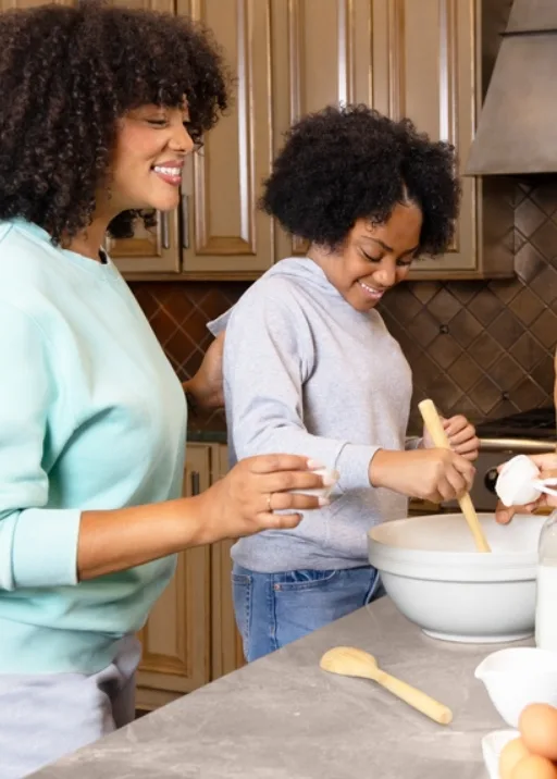 Mom and daughter are in the kitchen baking! The kitchen has glossy, light color wooden cabinets with a dark brown tile back splash. The island they are working on is a grey concreate color. Mom is wearing grey sweat shorts with a light, baby blue crew neck sweat shirt. She has her hand on her daughters back while the daughter is stirring something in a mixing bowl. The daughter is wearing jeans and a grey hoodie sweatshirt. They are both smiling and enjoying this bonding moment.