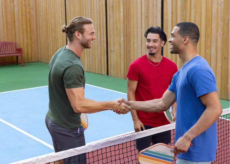 Three men at a backyard tennis court with a wooden fence. Two of then men are shaking hands on either side of a tennis net. The first man is in grey shorts and a green t-shirt. He is shaking hands with a man in a blue t-shirt holding a pickle ball racket. There is a man in a red crew neck t-shirt looking at them and smiling.