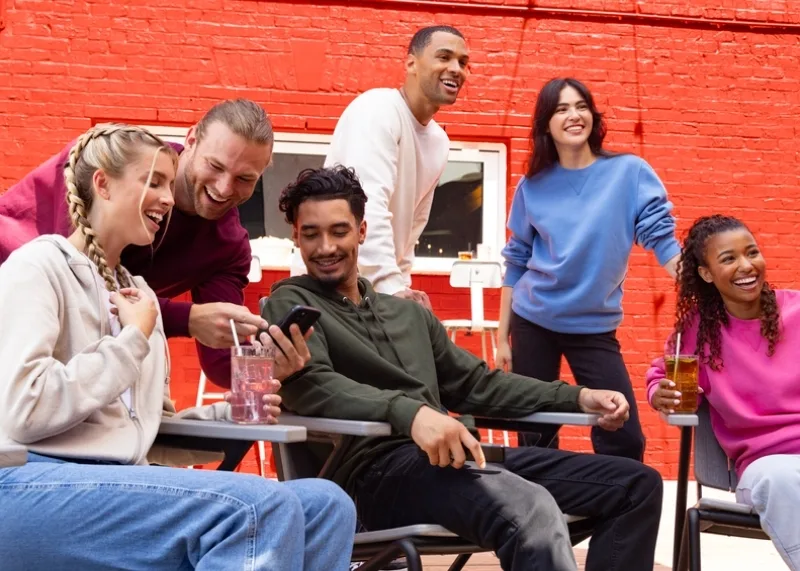 Six adults are shown gathered around outside, some are sitting in lawn chairs others are standing around. There is a red brick wall behind them. Theres a grouping of a women and two men chatting and laughing while looking at a phone. The women is sitting down and is in jeans and a cream zip up hoodie. The first man is standing up leaning down showing his phone and is wearing a maroon fleece crew neck sweatshirt. The man sitting down is in black jeans and a dark forest green hoodie. The next grouping of people are composed on one man and two women. The man and one women are standing while one women is sitting. All three are looking out in the distance. The man standing is wearing a white long sleeve crew neck shirt. The women standing is wearing black jeans and a light blue crew neck sweat shirt. The women sitting has on grey sweatpants and a bright pink crew neck sweat shit.