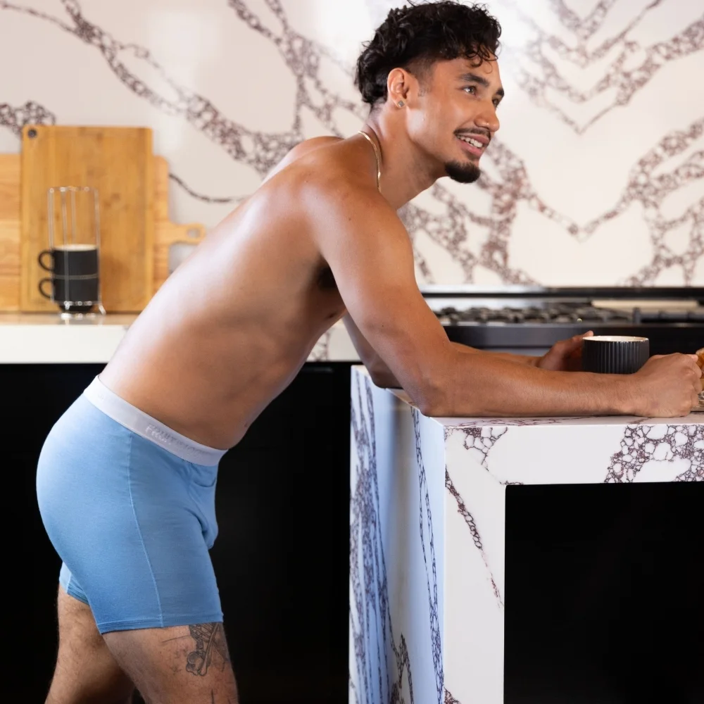 Male shirtless in baby blue boxer briefs leaning against kitchen counter. Behind him is the kitchen wall with a counter. The wall and counter are both made of what looks to be a slab of granite that is a cream color with these very heavy and thick light brown lines running all throughout it very randomly. Yes, there's granite on the wall. We only choose the fanciest of places to do photoshoots at! On the countertop you see a wooden cutting board leaning up against the wall and a stack of two fancy looking black coffee cups. There are black cabinets underneath. To the right of that is a black cooking range. In the foreground is a kitchen island that has a waterfall edge and its made of the same fancy granite looking stuff. The star of the photo is a well-tanned man with dark curly hair and a goatee and mustache combo. He isn't wearing a shirt but he is happy and smiling because he is wearing Fruit of the Loom boxer briefs in a light blue color with a greyish waistband. He is looking off to the side of the picture smiling at someone that we cannot see. 