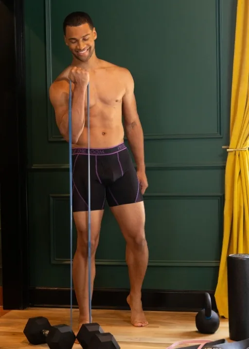 It's time to workout… in our boxer briefs. No shoes, no shirt, no problem because these Men's 360 Stretch boxer briefs are all you need! The boxer briefs are black with purple sticking around the legs and fly area. The waistband is black with the same purple color stitching that reads Fruit of the Loom. The room he is working out in is a light brown wooden floor with a dark green wall that has fancy woodwork on it. There is a mustard yellow curtain slightly out of frame. The guy is pulling up a stretchy resistance band. On the floor are various dumbbells and kettlebells.