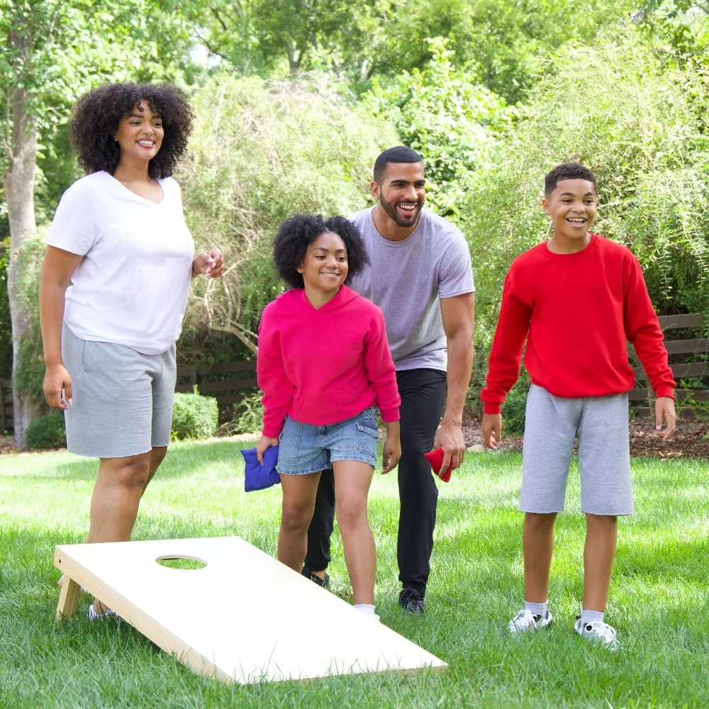 Its a bight, sunny day outside and the family decided to play cornhole on the front lawn. Mom is wearing grey fleece shorts with a white v neck t-shirt. She is watching while sister attempts to throw a blue cornhole bag. Sister is wearing jean shorts and a bight, pink hoodie. Dad is behind sister waiting for his turn with his red cornhole bag. Dad is wearing black joggers and a grey crew neck t-shirt. Brother is cheering them on from the side wearing grey fleece shorts and a red fleece crew neck sweatshirt.