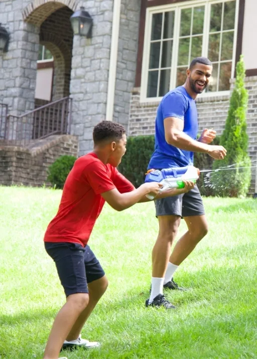 Outdoor summer fun! Dad and son playing with a water gun. The son is wearing dark navy shorts and a red crew neck t shirt. He has the water gun and is spraying at dad who is running away smiling. Dad has on the classic dad sneakers with white socks that go up high on his leg. He also has on grey fleece shorts and a soft, blue crew neck t shirt.