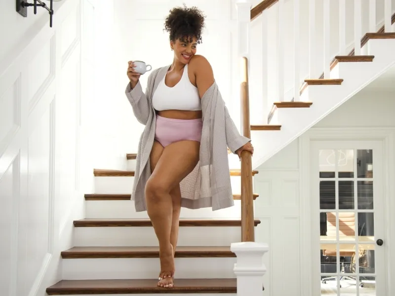 women wearing fruit of the loom pink underwear and white bra standing on a staircase with a cup of coffee. She also has khaki colored cardigan draped around her arms and shoulders.