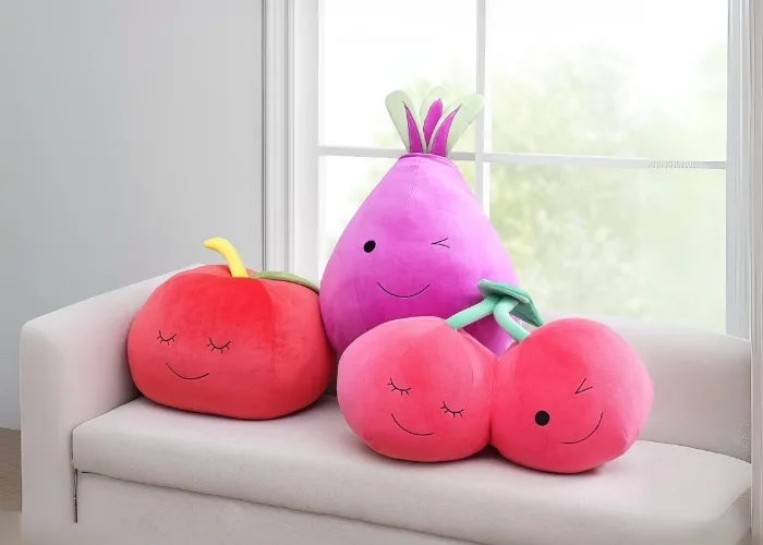 This photo is of three pillows sitting on a bench in front of a window. Since this is Fruit of the Loom, all three pillows are fruit shaped! The first is a red apple that is smiling in its sleep. The second is a purple berry that has three pieces of hair and its face is winking at you. The last pillow is pink twin cherrys with green stems connecting the two. The first of the cherries is smiling in its sleep the second is winking at you because you know, its perfectly normal for fruit to have faces.