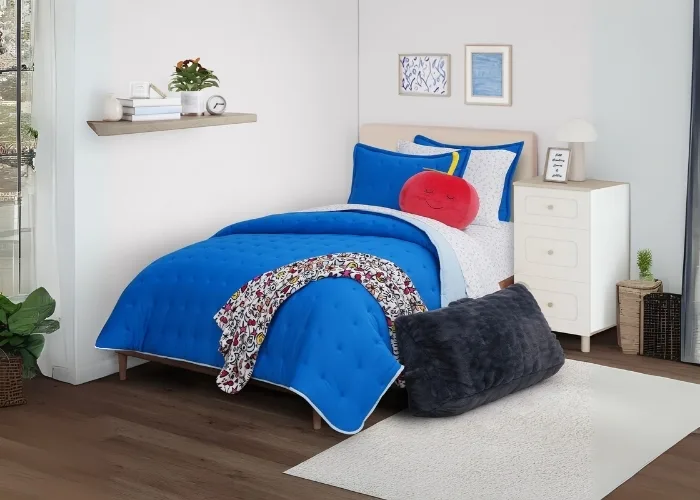 This is a bedroom scene. The bed is a tan color headboard. Next to it is a night stand. On the wall next to it is a shelf. The bedding on the bed is the main focal point of the photo. The bedding is bright blue. There is a throw blanket draped over a corner that has a fruit design on it. Also on the bed is a throw pillow that looks like an apple, just like in the fruit of the loom logo! Except, this apple has a face on it that looks like its smiling in its sleep. On the floor is a white rug and a black wedge pillow that is large enough that you would use it to lean against.