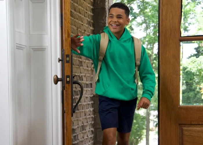 Back to school time! Brother is coming home from school, entering through the wooden front door. He is as happy as can be while wearing navy fleece shorts and a kelly green hoodie with his tan backpack on.