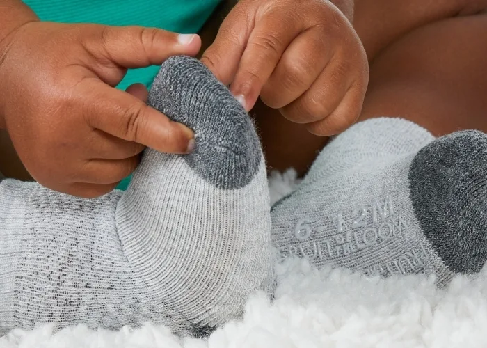 A close up photo of a baby where you see its hands playing with a pair of grey baby socks that have 6 to 12 months written on the bottom of the sock in a grippy material.