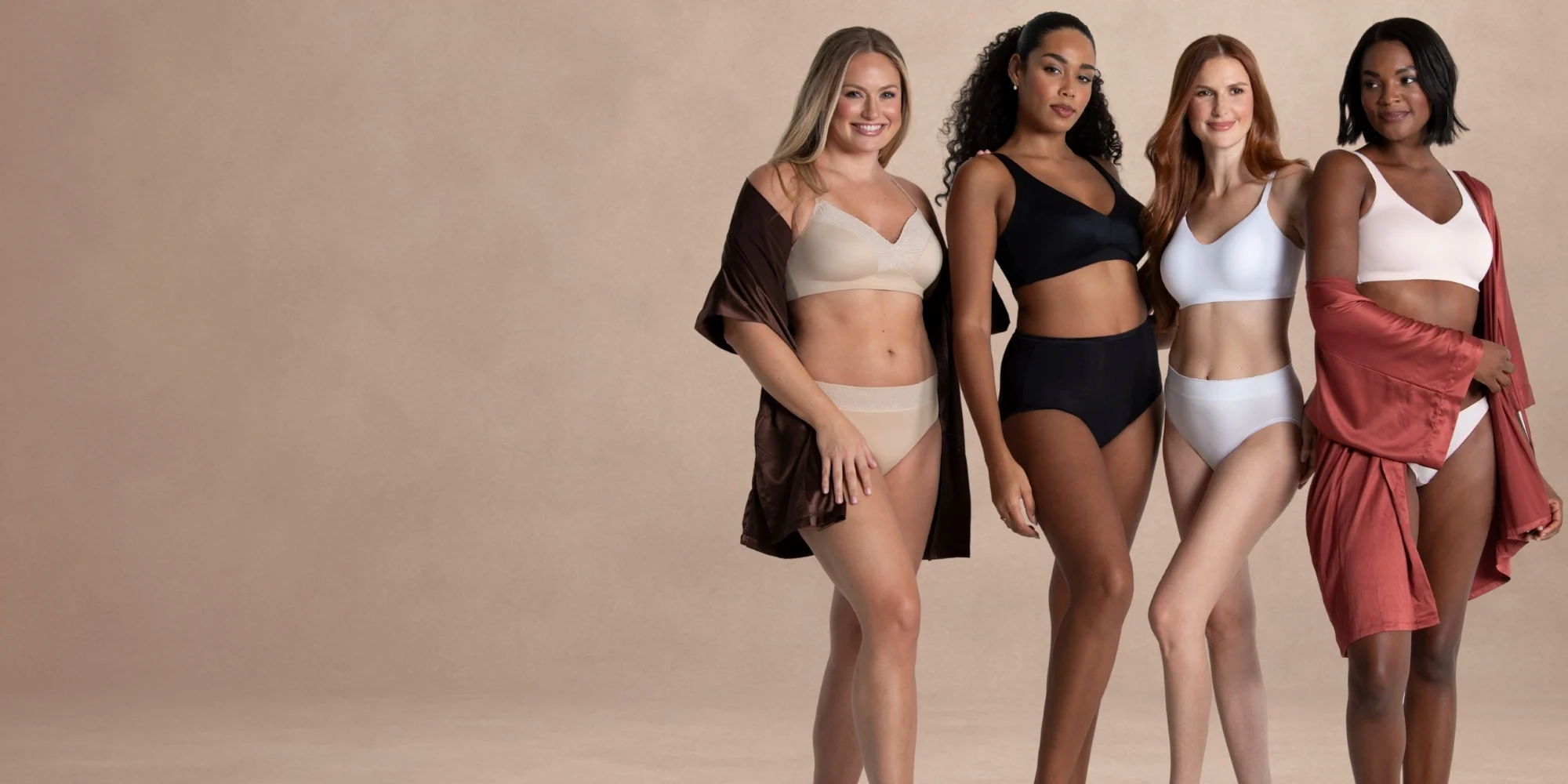 Four diverse women wearing neutral-toned bras and panties from Vanity Fair Lingerie stand confidently against a soft beige textured backdrop. They are smiling and relaxed, showcasing the comfort and fit of the lingerie styles.