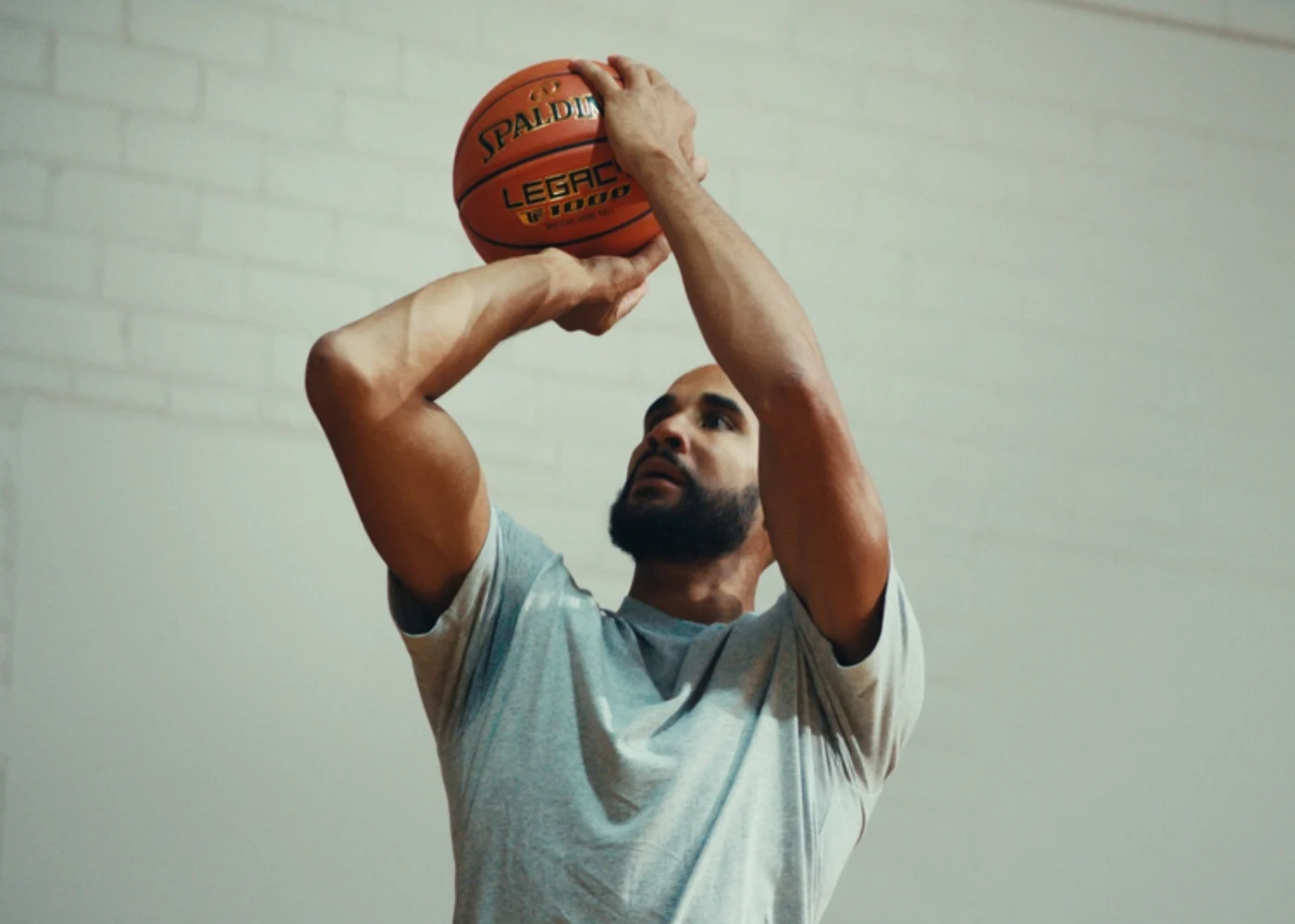 Basketball player in grey shirt preparing to shoot Spalding Legacy TF-1000 indoors against white brick wall.