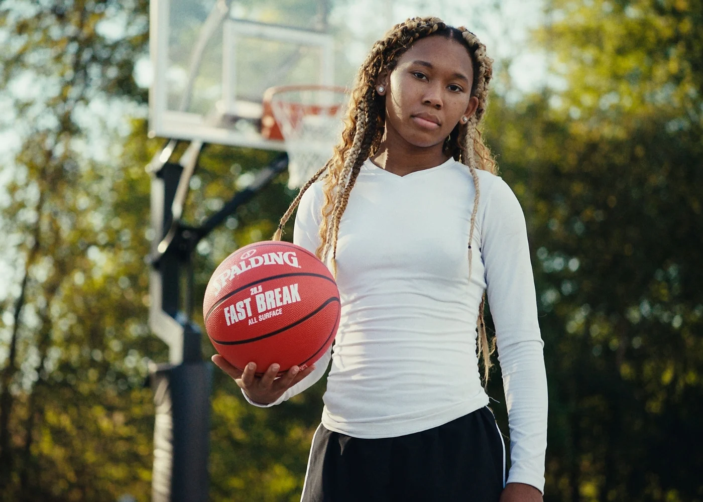 Female holding Spalding Fast Break basketball on outdoor court with hoop and trees in background.
