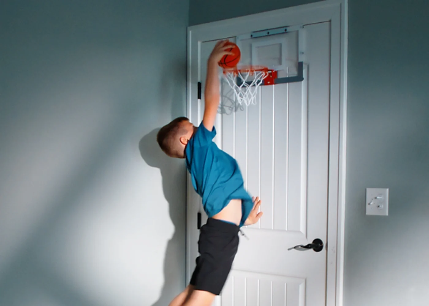 Young boy dunking basketball on indoor mini hoop mounted to white door in home setting.