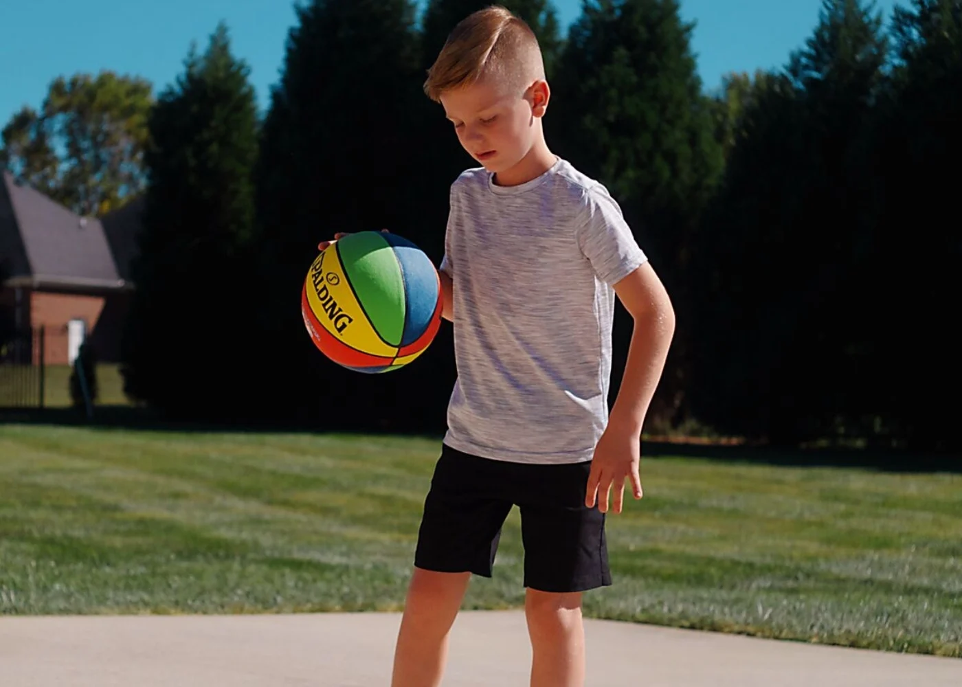 Child holding colorful basketball on outdoor court with green grass and trees, wearing gray t-shirt and black shorts.
