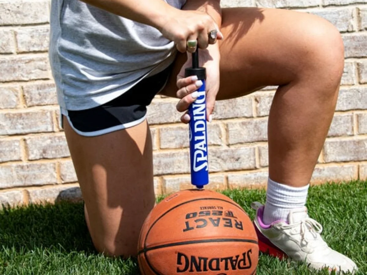 Person inflating a Spalding React basketball with a hand pump outdoors on grass, demonstrating basketball accessories and equipment for recreational play.