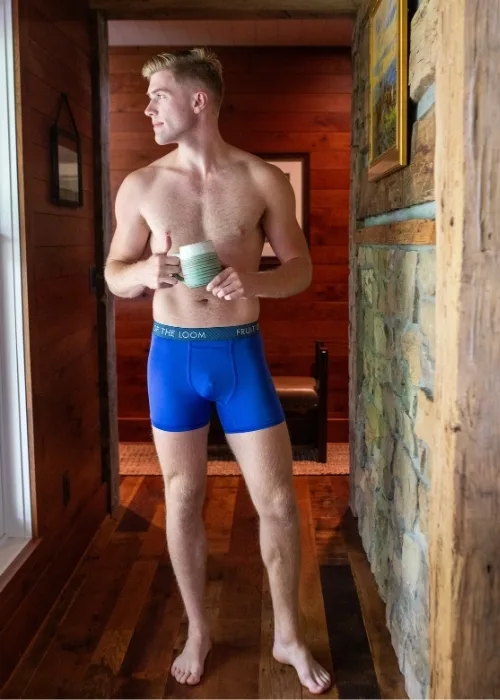 Shirtless man wearing blue boxer briefs standing and looking out a window while holding a coffee mug.