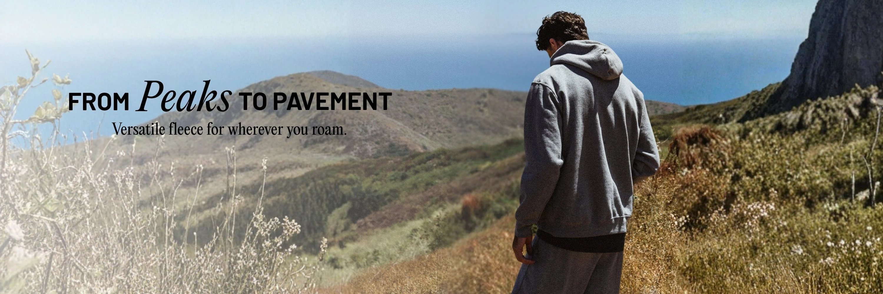 Man wearing Russell athletic hoodie and sweatpants standing on a mountain range looking out over the landscape. With text overlay: From Peaks to Pavement Versatile fleece for wherever you roam.