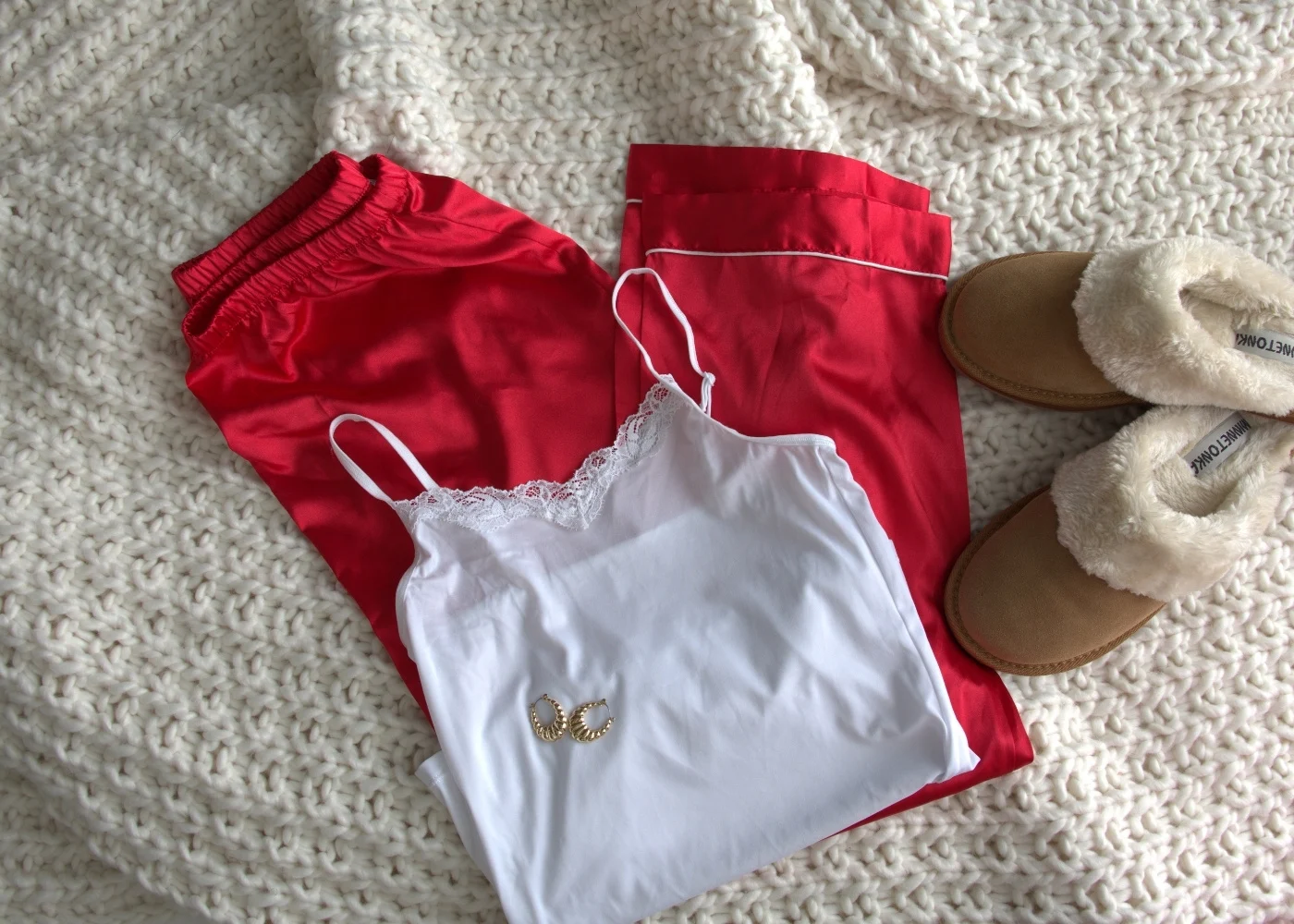 A white lace-trim camisole paired with vibrant red satin pajama pants and tan fuzzy slippers, all arranged on a chunky cream knit blanket. Gold hoop earrings complete the festive and cozy look.