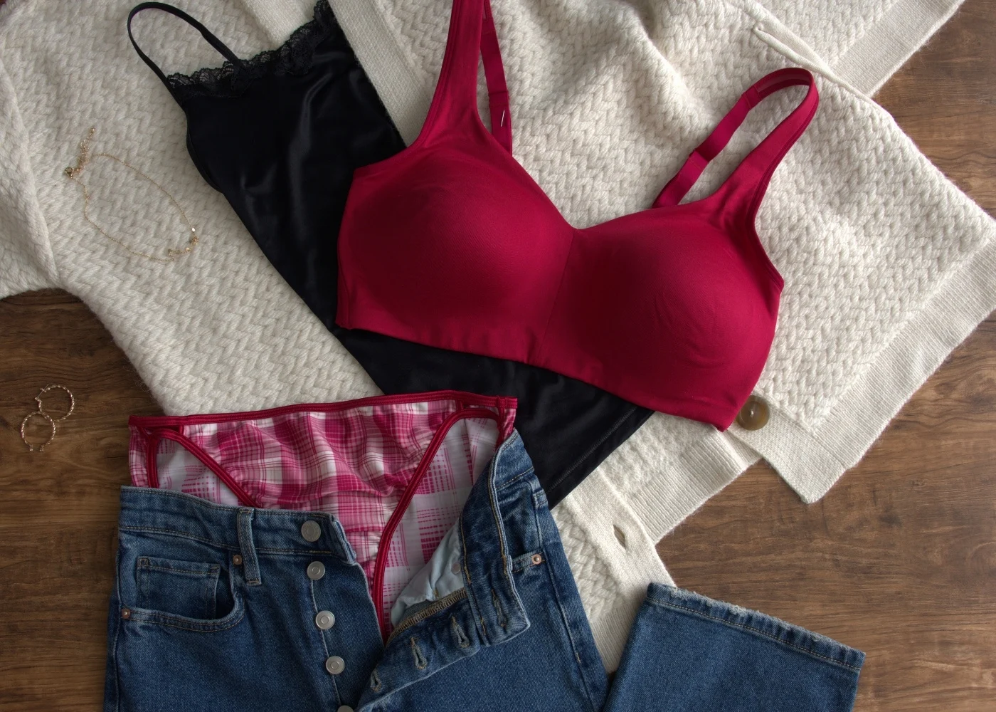 A flat lay featuring a bright red bra, a black camisole, and a pair of plaid panties arranged on a cream knit sweater. A pair of blue jeans is partially folded at the bottom of the frame, creating a cozy and casual styling theme.