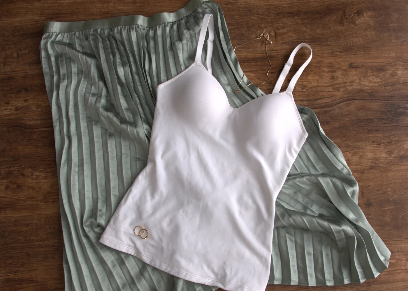 A white camisole with built-in support displayed on top of a soft, pleated sage green skirt. The setup includes delicate gold jewelry, adding an elegant and minimal aesthetic.
