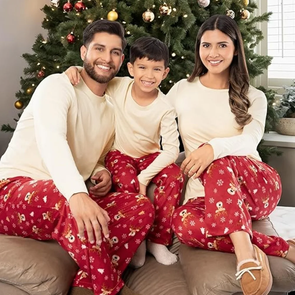Three people sitting together in front of a decorated Christmas tree, wearing matching Fruit of the Loom holiday-themed pajamas with cream-colored tops and red pants featuring gingerbread and snowflake patterns.