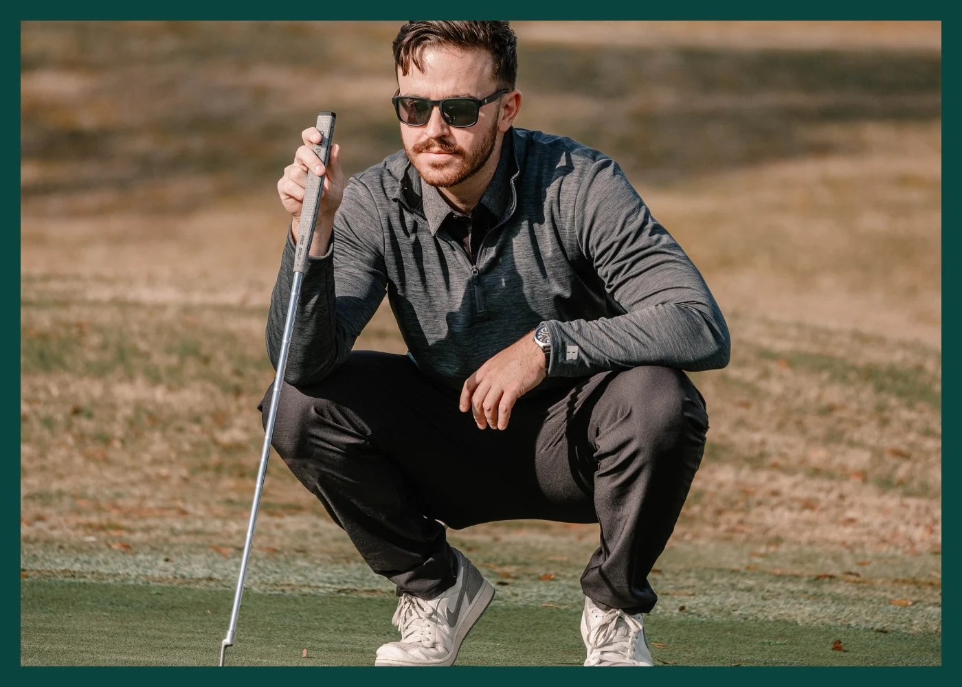 Male model wearin ggrey  polo and grey pants, holding a putter on a green.