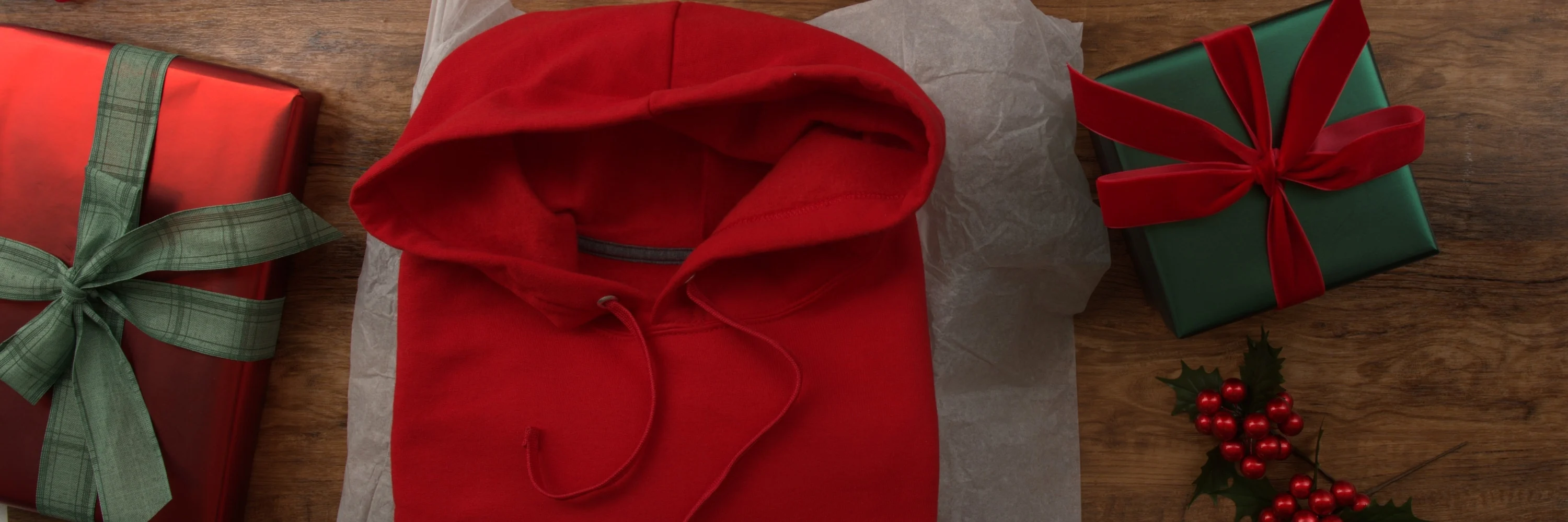 Christmas gift idea: red hoodie with holiday wrapping paper, presents, and ornaments on rustic wood.