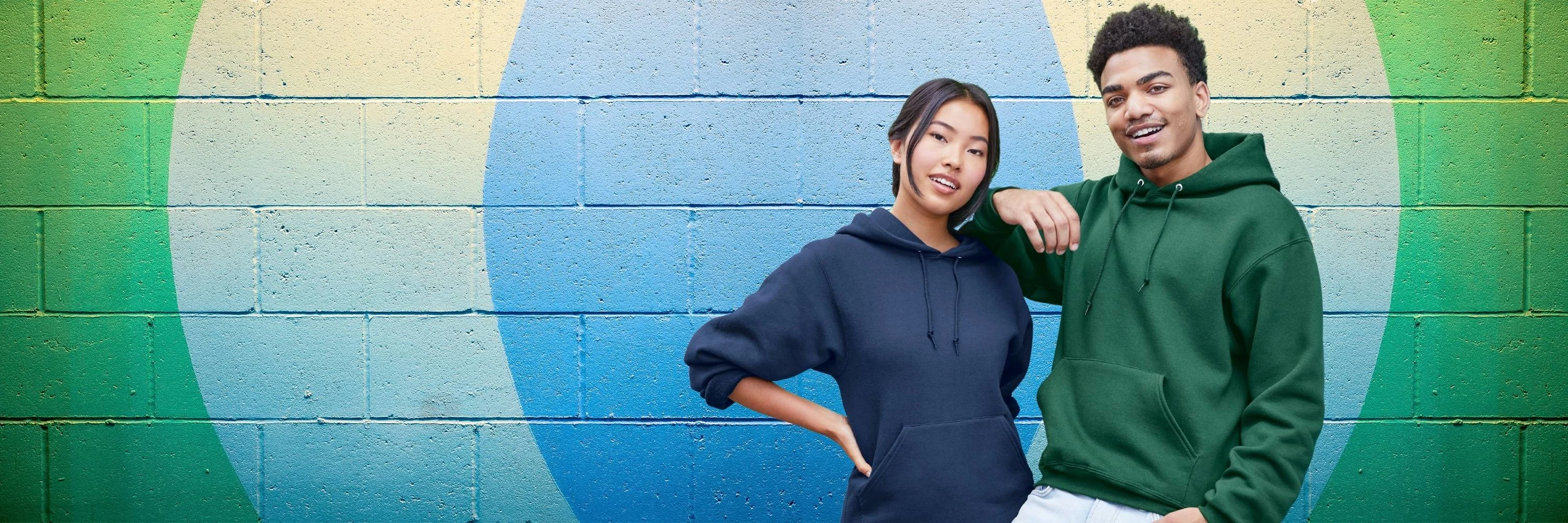 Man and woman posing in front of a brick mural wearing fruit of the loom navy blue  and forest green fleece hoodies 