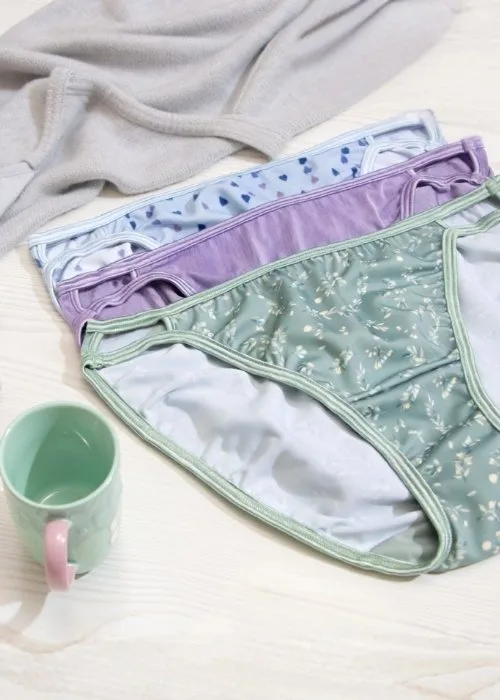 Three pairs of women’s panties in assorted colors and prints are neatly arranged on a light wood surface, with a soft gray sweater and a ceramic mug nearby, creating a warm, cozy flat‑lay scene.