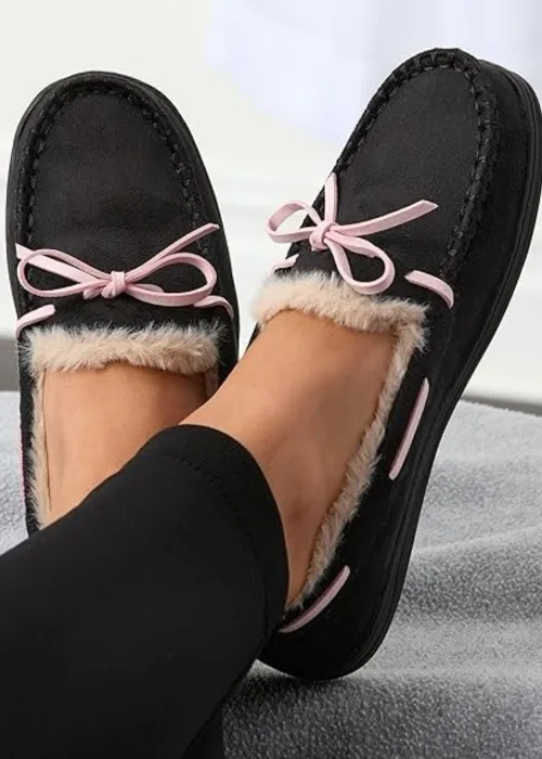 Women’s black moccasin-style slippers with pink bows and faux fur lining, worn with leggings and shown on a soft grey surface to highlight cozy indoor footwear for cold weather.