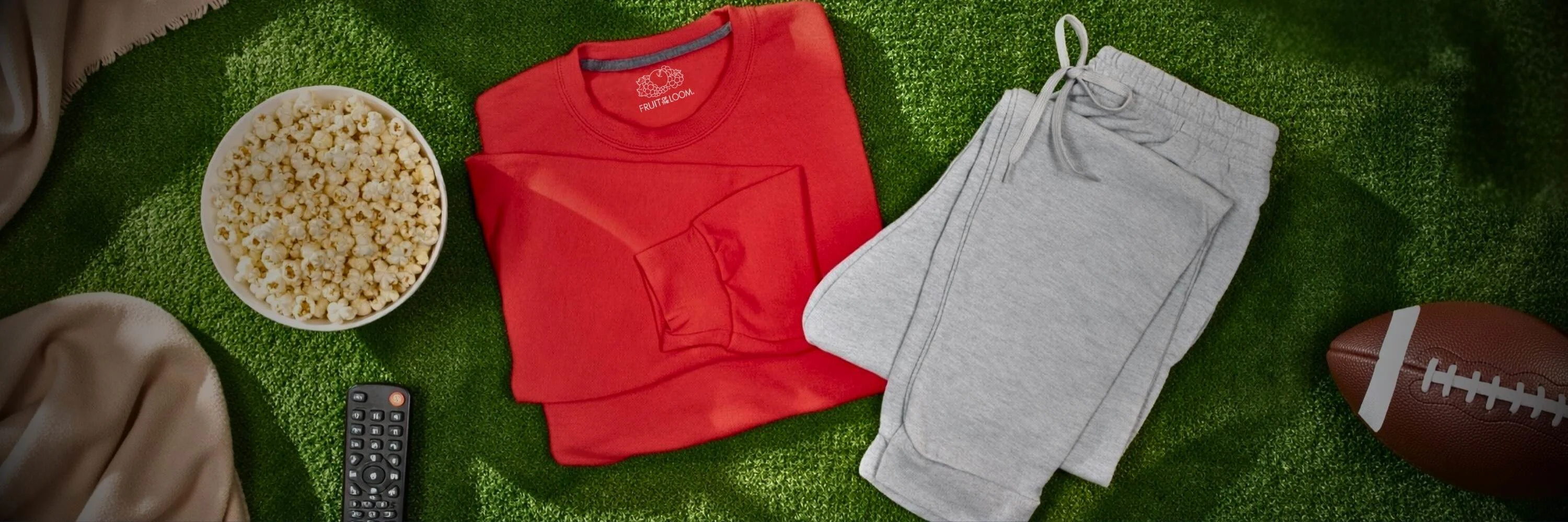 Folded red sweatshirt and light gray fleece joggers arranged on green turf with a bowl of popcorn, TV remote, beige blanket, and a football.”