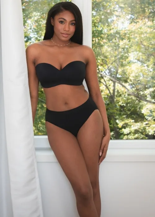 Model wearing the new beyond comfort wireless strapless bra in midnight black posing by a window in matching midnight black hicut panties
