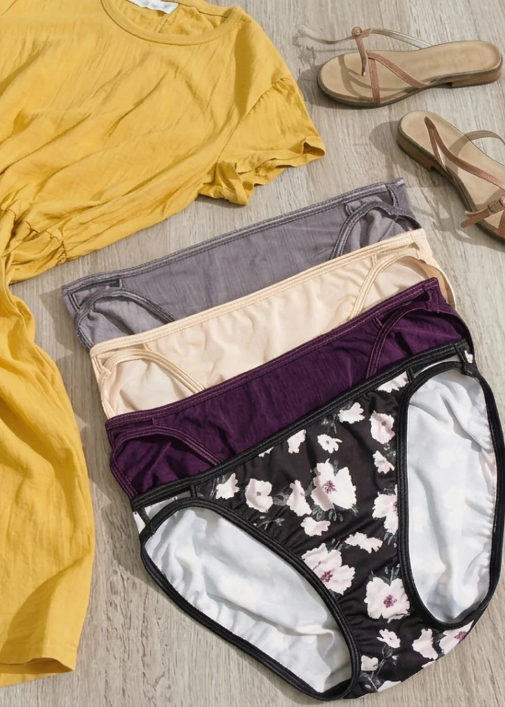 Assorted women’s panties in neutral shades styled with a light summer dress, hat, sunglasses, woven bag, and sandals on a linen background.