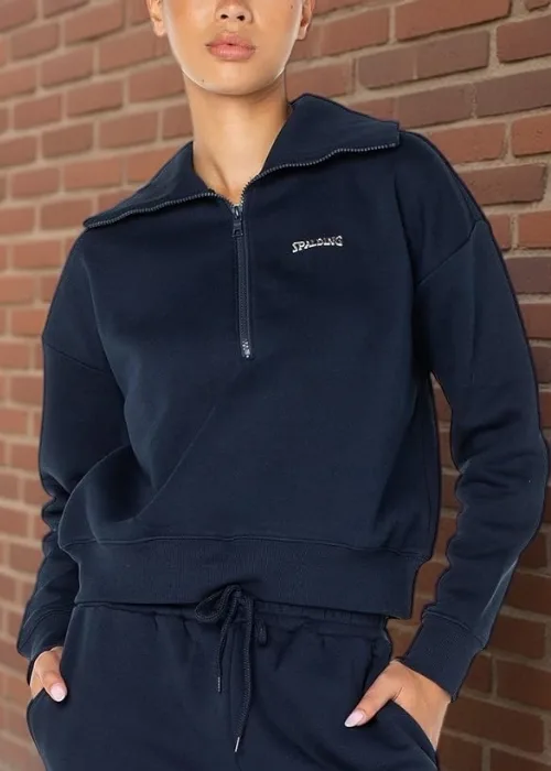 Model wearing a navy Spalding quarter-zip pullover and matching joggers standing against a brick wall along a city sidewalk with parked cars and pedestrians in the background.