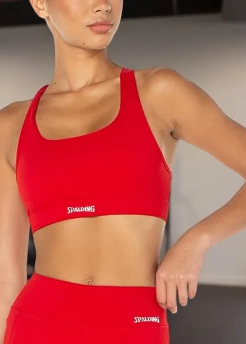 Model wearing a red Spalding sports bra and matching high-waisted leggings standing in a modern gym with mirrors and a dumbbell rack in the background.