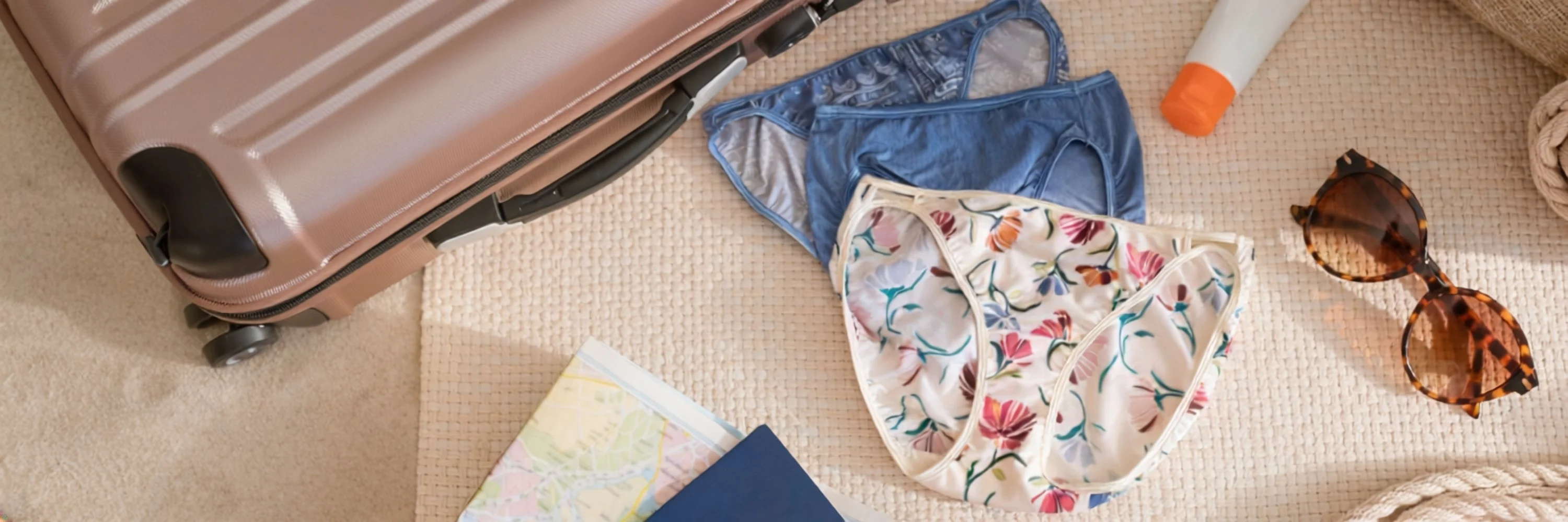 A set of blue and floral women’s underwear arranged beside a suitcase, sunglasses, sunscreen, and a map, evoking a summer travel packing scene.