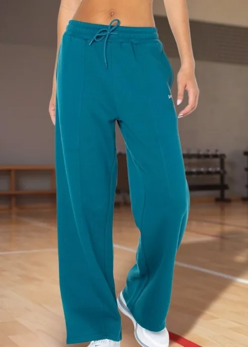 Model wearing teal Spalding sweatpants and white sneakers standing on an indoor basketball court with wooden flooring, weight racks, and a ball cart in the background.