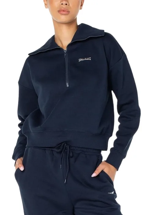 Model wearing a navy Spalding quarter-zip pullover and matching joggers standing against a brick wall along a city sidewalk with parked cars and pedestrians in the background.