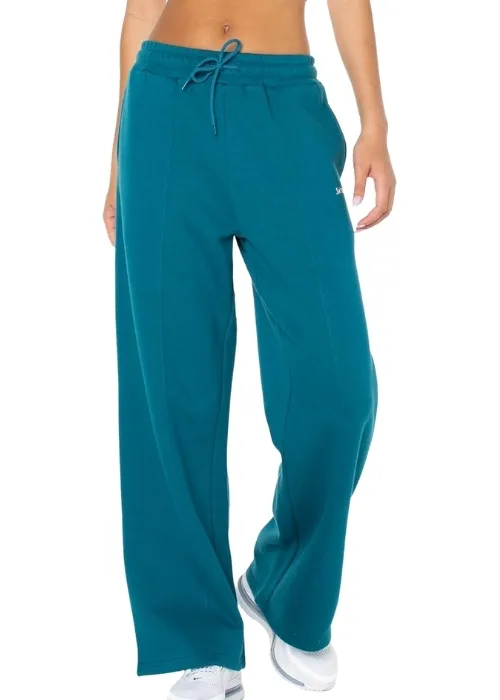 Model wearing teal Spalding sweatpants and white sneakers standing on an indoor basketball court with wooden flooring, weight racks, and a ball cart in the background.
