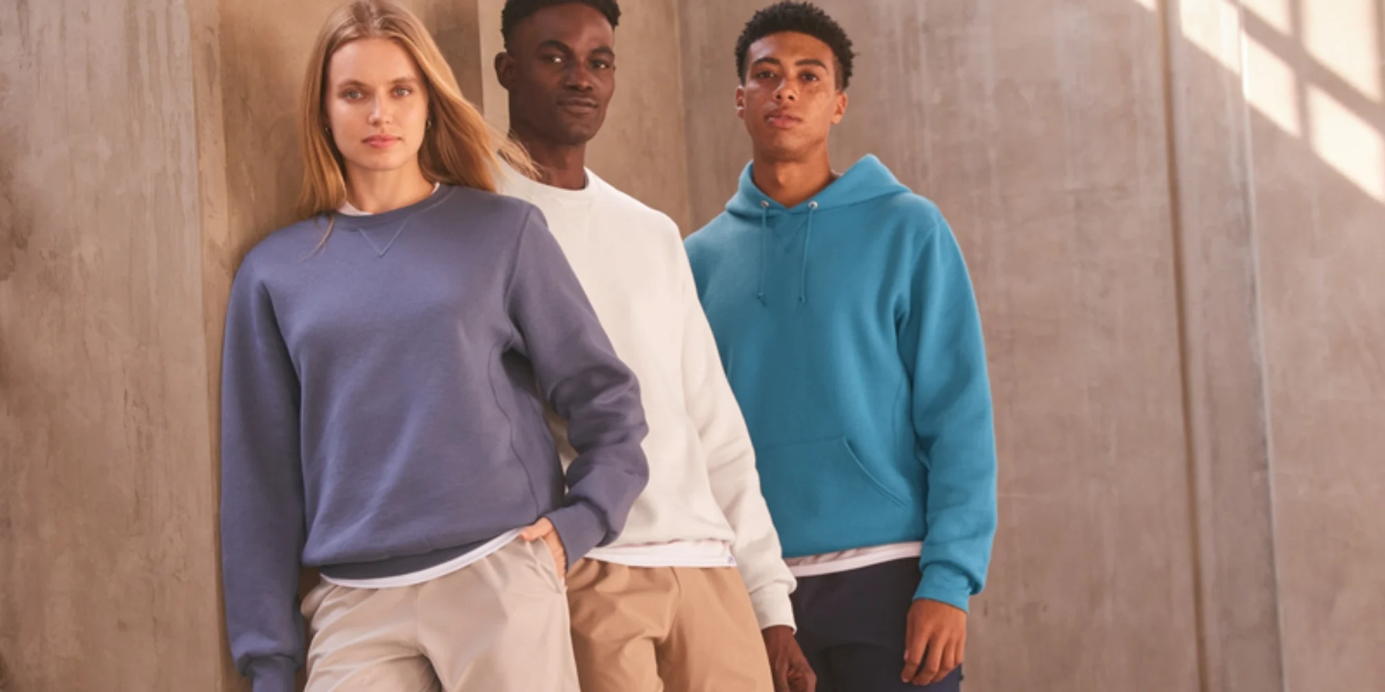 Three people wearing Russell Athletic fleece apparel, including a blue hoodie, crewneck sweatshirt, and comfortable everyday essentials, posed against a concrete wall in natural sunlight.